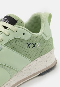 Sneaker verde chiaro in suede perforato, logo "XX" cucito in bianco e nero, suola texturizzata macchiata di punti marroni e colletto imbottito.