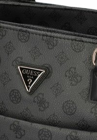 Borsa nera in tessuto con un motivo sottile, caratterizzata da un logo metallico triangolare con "GUESS U.S.A." e un punto interrogativo.