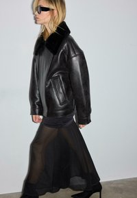 Blonde Frau in übergroßer schwarzer Lederjacke mit Pelzkragen, durchscheinendem schwarzem Maxirock, schwarzen hochhackigen Stiefeln und dunkler Sonnenbrille geht nach links.