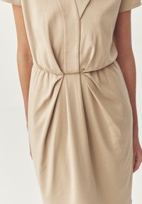Abito beige con un tessuto morbido e liscio. Presenta un dettaglio a torsione frontale in vita e maniche corte. Design semplice ed elegante.