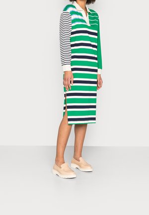 Vestido de rayas con colores verde, azul marino y blanco, mangas largas, cuello y aberturas laterales. Combinado con zapatos tipo mocasín de color claro.