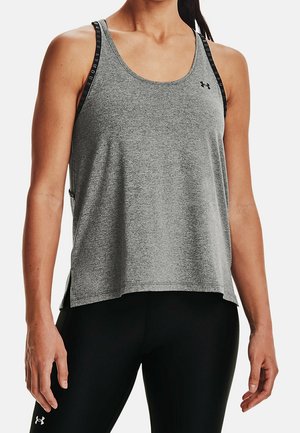 Kvinde iklædt grå ærmeløs sportstop og sorte leggings med synlige mærkelogoer på stropperne og benet.