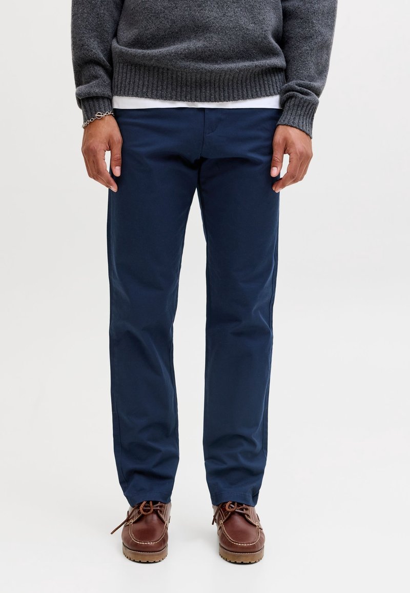 Homme portant un pantalon bleu marine, des chaussures en cuir marron, un pull gris et un sous-chemise blanche sur un fond clair uni.