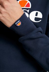 Sudadera azul marino con puños acanalados. Presenta un gran logo blanco de "ellesse" y un parche de logo colorido en el puño. Textura suave.