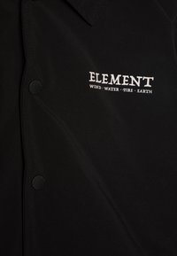 Crna majica s glatkom teksturom, koja ima kopčanje na dugmad i logo koji glasi "ELEMENT" s natpisom "VJETAR, VODA, VATRU, ZEMLJA" ispod.