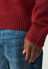 Rdzawy, dziany sweter z prążkowanymi mankietami, w zestawie z niebieskimi dżinsami. Sweter ma teksturalne wykończenie z subtelnymi kolorowymi nitkami.
