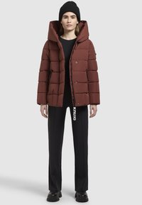 Dunkelrote Pufferjacke mit großer Kapuze, gestepptem Design, vorderem Druckverschluss und Seitentaschen. Getragen mit schwarzen Hosen und einer Mütze.