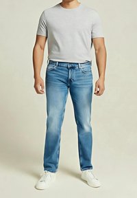 Mężczyzna ubrany w jasnoszarą koszulę z krótkim rękawem, niebieskie dżinsy i białe trampki, stojący na tle jednolitego, jasnego tła.