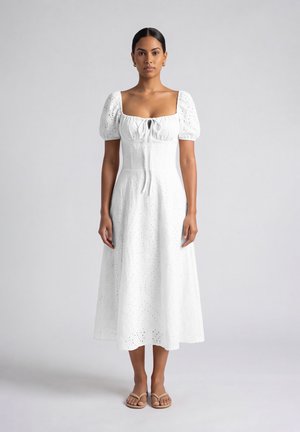 Vrouw staand in een witte midi-jurk met oogjes, pofmouwen en beige sandalen tegen een effen lichtgrijze achtergrond.
