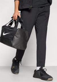 Μαύρη τσάντα duffel Nike με μπροστινή τσέπη με φερμουάρ, μαύρο αθλητικό παντελόνι και μαύρα αθλητικά παπούτσια με πορτοκαλί λεπτομέρειες. Υλικό: συνθετικό, με λεία υφή.