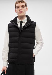 Μαύρο μπουφάν αμάνικο με ψηλό γιακά και φερμουάρ μπροστά. Διαθέτει καπιτονέ σχέδιο και διακριτικό λογότυπο. Φοριέται πάνω από λευκό πουκάμισο και γραβάτα.