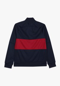 Tummansininen takki, jossa on silmiinpistävä punainen vaaka-aita selässä. Takki on valmistettu sileästä, kevyestä kankaasta ja siinä on pystykaulus.