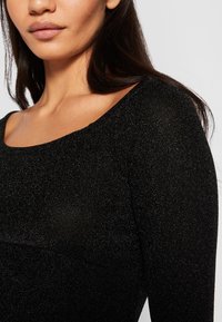 Musta, istuva pitkähiha paita, jossa on leveä pääntie ja teksturoitu, kimaltava kangas, joka heijastaa valoa hienovaraisilla kimalteilla.