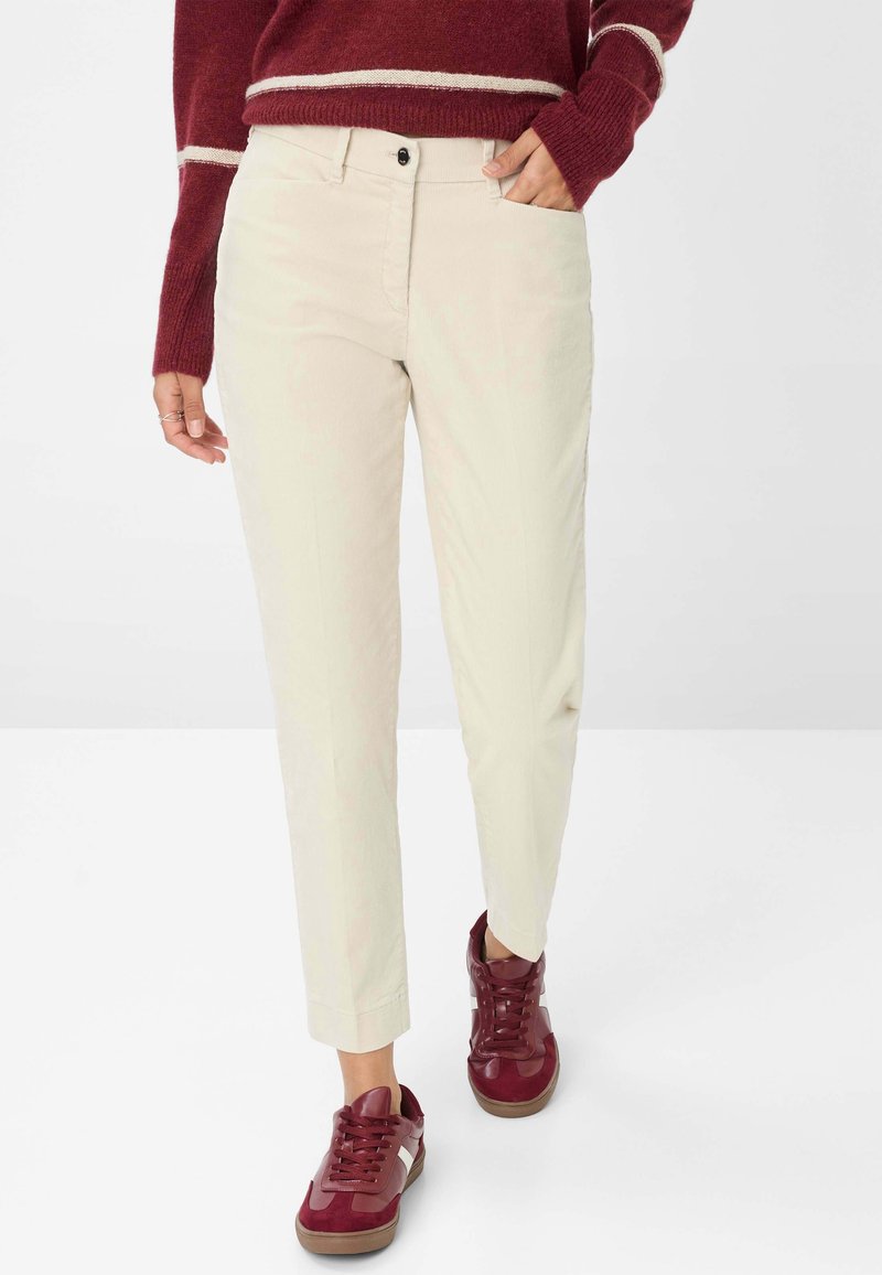 Femme portant un pantalon beige droit, un pull bordeaux à rayures blanches et des baskets bordeaux, debout avec une main dans la poche.