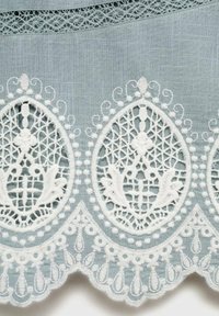 Tissu bleu clair avec bordure en dentelle blanche brodée complexe présentant des motifs en forme de goutte et des bords festonnés. Les détails incluent des éléments en relief.