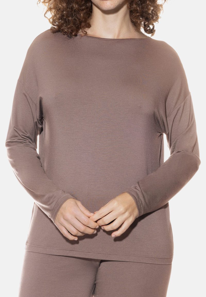 Langsleeve top in taupe, gemaakt van zachte stof. heeft een wijde hals en een losse pasvorm. Handen zijn in elkaar geklapt op de taille.