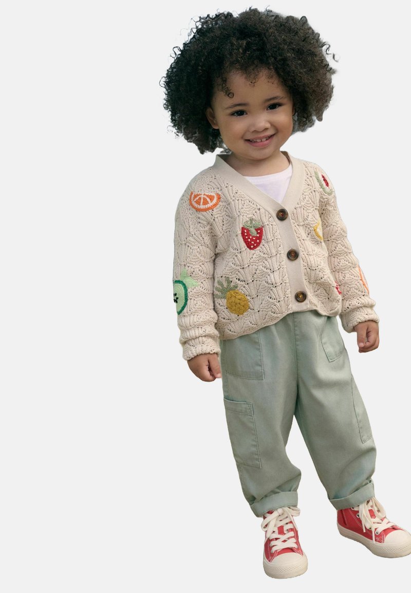 Cárdigan beige de punto con bordado de frutas, botones marrones y camisa blanca, combinado con pantalones verdes y zapatillas rojas con suelas blancas.