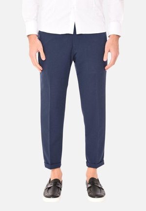 Pantaloni blu navy con un design slim fit, orli arricciati, texture liscia e due tasche frontali, abbinati a scarpe nere slip-on.