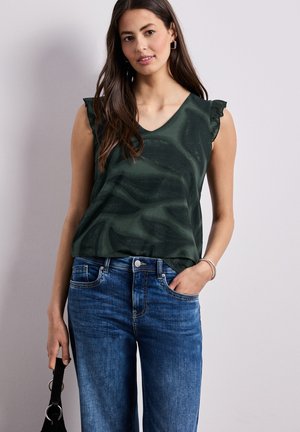 Kvinde iført en mørkegrøn ærmeløs bluse med flæser på skuldrene og blå jeans, der holder en sort håndtaske i den ene hånd.