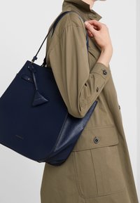 Navy nahkaämpäri laukku tekstuuripinnalla, jossa on säädettävä hihna ja pieni merkkikoriste. Päälle puettuna vaaleanoliivinvihreän vaatteen kanssa.