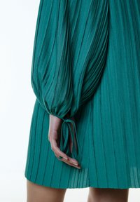 Teal jurk met verticale plooien, voorzien van volumineuze lange mouwen en een gestrikt detail bij de manchet. Soepele, gestructureerde stof.