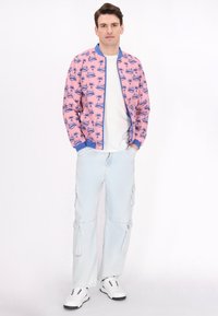 Giacca bomber rosa con stampa di palme, dettagli blu, t-shirt bianca, pantaloni cargo azzurri e sneakers bianche con suole nere.