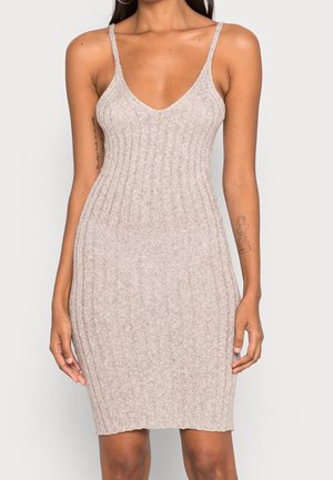 Vrouw die een mouwloze, beige geribbelde gebreide jurk met spaghettibandjes draagt, staand met haar armen ontspannen langs haar zij op een effen achtergrond.