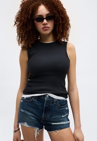 Νεαρή γυναίκα με σγουρά μαλλιά που φοράει μαύρο μανίκι τοπ, σκούρα γυαλιά ηλίου, σκισμένο τζιν σορτς και βραχιόλι, με φόντο λευκό.