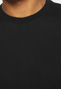 Camiseta de algodón negra con cuello redondo y mangas cortas. Presenta una textura sutil y un pequeño logo bordado en el pecho.