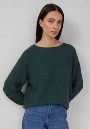 Frau mit langen dunklen Haaren, die einen dunkelgrünen strukturierten Strickpullover und blaue Jeans trägt, steht vor einem schlichten grauen Hintergrund.