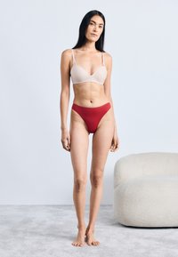 Lichtbeige beha met dunne bandjes, gecombineerd met rode high-waisted slipjes. Model staat op een gestructureerde lichtgrijze vloer, naast een zachte fauteuil.