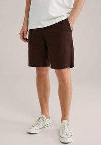 Pantalones cortos de algodón marrón con un corte recto, bolsillos laterales y un dobladillo limpio, combinados con zapatillas blancas con detalles en rojo y azul marino.