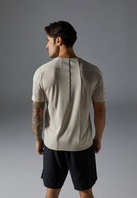 T-shirt de sport beige clair avec un dos texturé, manches courtes et une rayure verticale grise. Associé à un short noir, mettant en avant un design ajusté.