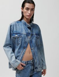 Γυναίκα με βρεγμένα μαλλιά, φοράει ανοιχτό γαλάζιο τζιν μπουφάν χωρίς κουμπιά πάνω από τζιν παντελόνι που ταιριάζει, με το ένα χέρι στην τσέπη, μπροστά σε απλό φόντο.