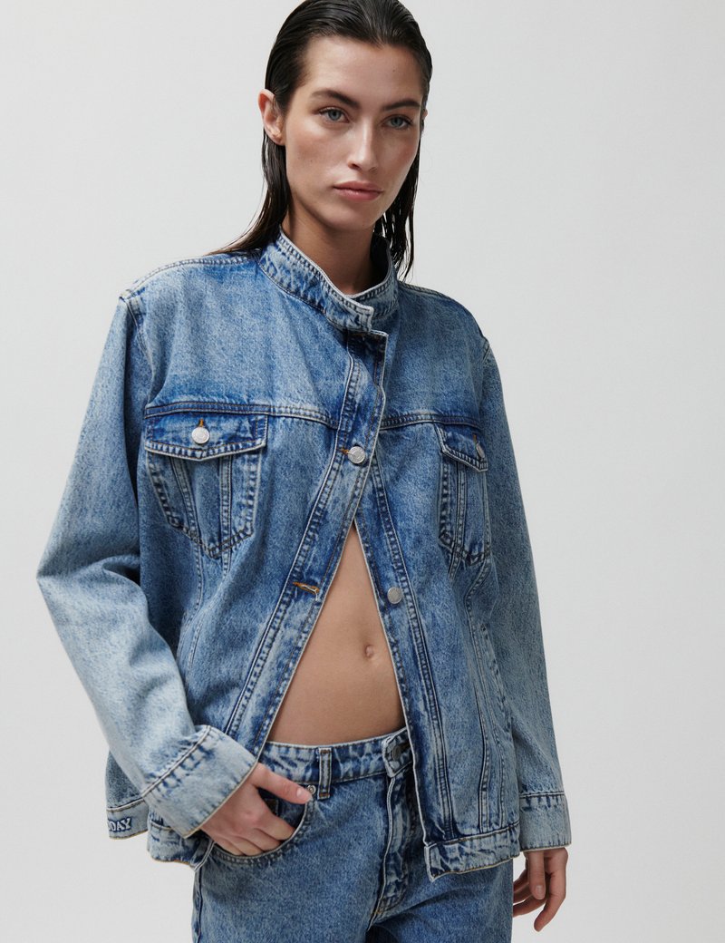 Γυναίκα με βρεγμένα μαλλιά, φοράει ανοιχτό γαλάζιο τζιν μπουφάν χωρίς κουμ πιά πάνω από τζιν παντελόνι που ταιριάζει, με το ένα χέρι στην τσέπη, μπροστά σε απλό φόντο.