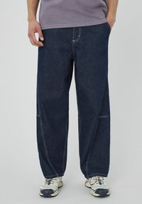 Pantalons en denim bleu foncé à jambes larges, avec des coutures blanches contrastantes et des poches latérales, associés à des chaussures de sport de couleur claire.