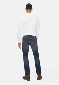 Jean en denim bleu foncé avec une légère extensibilité, coupe droite, et des détails de couture discrets sur les poches arrière, associé à une chemise blanche à manches longues.