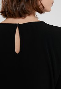 Zwarte blouse met een ronde halslijn, een sleutelgatopening aan de achterkant en een knoopsluiting. Gemaakt van een gladde, lichte stof.