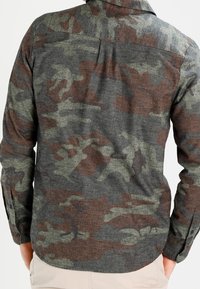 Chemise à manches longues camouflage en tissu gris, vert et marron ; possède un col boutonné, un empiècement dos simple et des poignets ajustables.