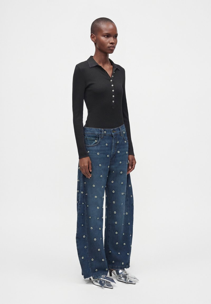Mannequin portant un top noir moulant à manches longues, un jean bleu à jambes larges avec des embellissements circulaires et des talons argentés pointus sur fond blanc.