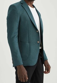 Blazer teal realizzato in tessuto testurizzato, con due bottoni, una tasca sul petto e polsini dettagliati, abbinato a una camicia bianca sotto.