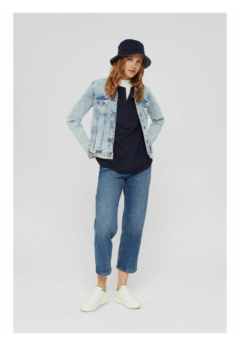 Frau mit einem marineblauen Bucket Hat, einer hellen Jeansjacke, einem marineblauen Hemd, blauen cropped Jeans und weißen Sneakers, die mit den Händen in den Jackentaschen steht.