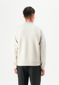 Suéter creme com um tricot texturizado, decote redondo e ombros caídos, combinado com calças cinza-escuras. Vista traseira, design sem costura.