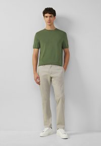 Groene shirt met korte mouwen van gebreid materiaal, gecombineerd met lichtbeige broeken en witte sneakers, tegen een effen grijze achtergrond.