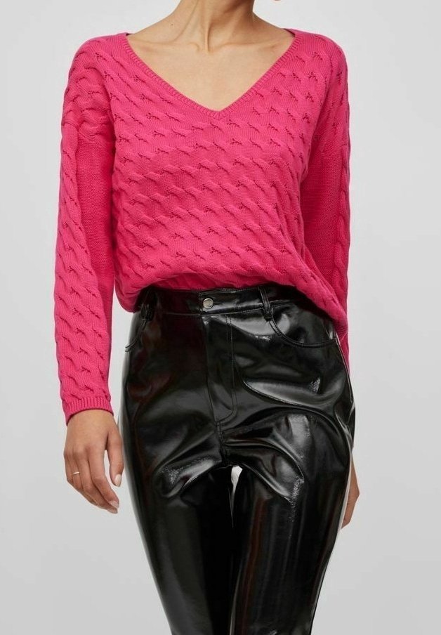 Femme portant un pull en maille texturé rose vif et un pantalon en vinyle noir brillant à taille haute, debout devant un fond clair uni.