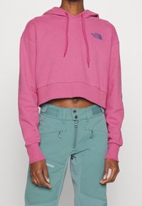 Sudadera con capucha recortada en color rosa brillante con dobladillo acanalado; presenta un logo azul en el pecho. Combinada con pantalones verdes con múltiples bolsillos.