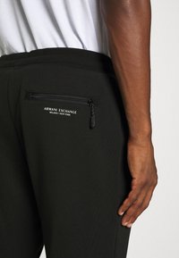 Crne trenirke s bočnim džepom na zip, s bijelim natpisom "Armani Exchange Milano/New York" i glatkom, teksturiranom tkaninom.