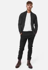Grijze ritscardigan over een witte T-shirt, zwarte slim-fit broek en bruine veterschoenen. Glad van structuur met een aansluitend ontwerp en minimale details.