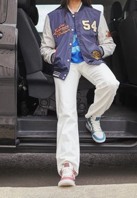 Blauw en grijs varsityjack met geborduurde details, witte wijde pijpen broek en meerkleurige hoge sneakers, gepositioneerd in een autodeur.