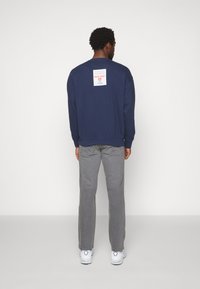 Marineblauwe sweatshirt met lange mouwen, geribde manchetten en een rechthoekig label op de achterkant. Gecombineerd met grijze jeans en witte sneakers.